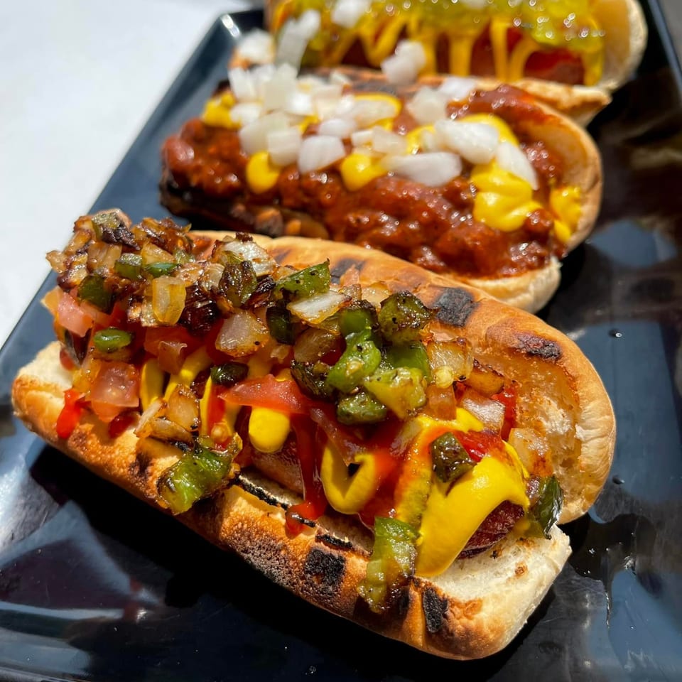 Three Aljoom BBQ hot dogs with various toppings, including chili, onions, and peppers.
