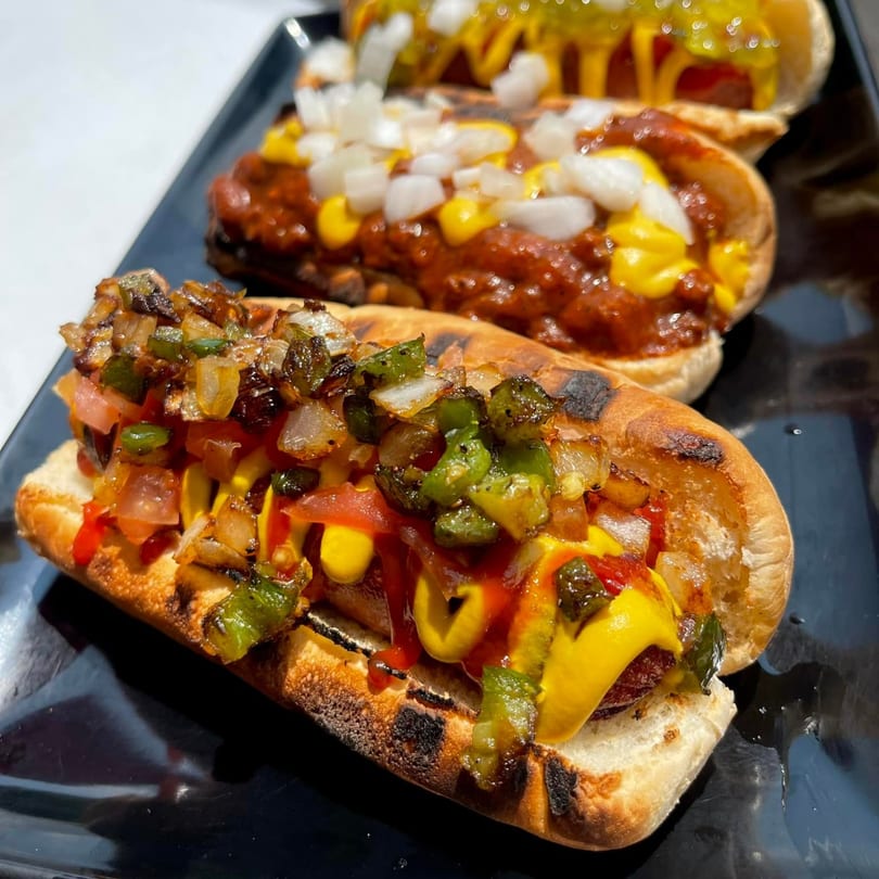 Three Aljoom BBQ hot dogs with various toppings, including chili, onions, and peppers.