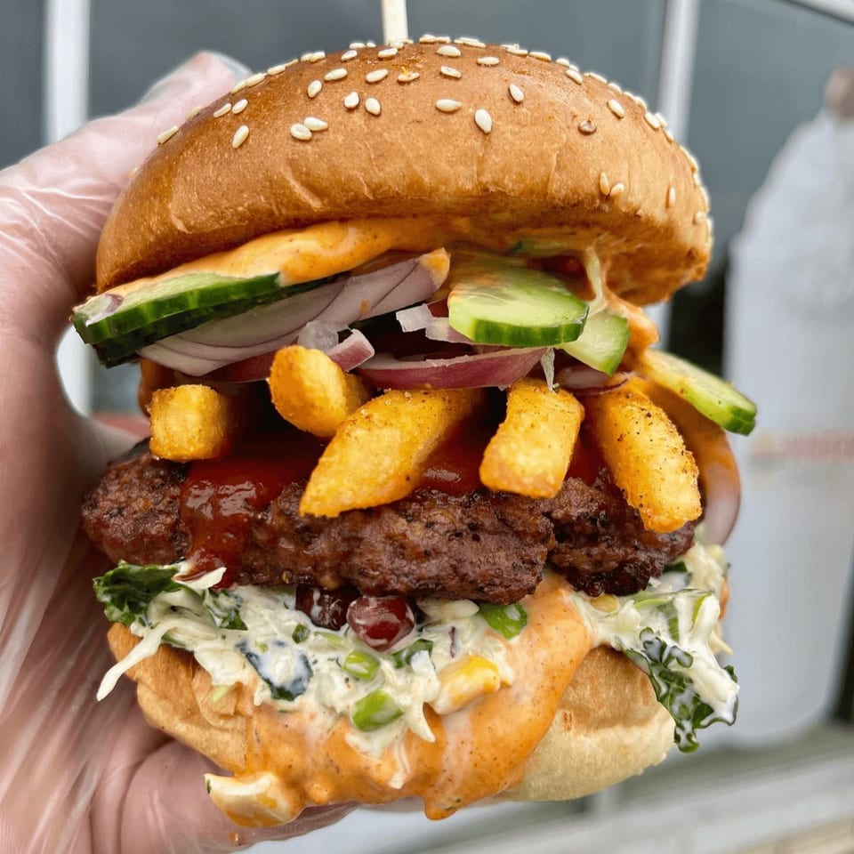 A hand in a clear glove holds a large Aljoom BBQ burger with fries, cucumbers, and red onions.